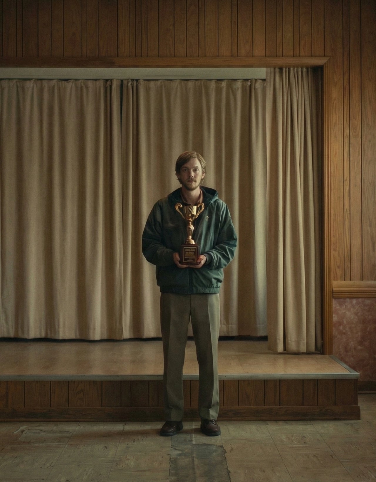 A young man stands alone in a wood-panelled community hall holding a small trophy. An empty podium stands to his left. Folding chairs line the wall to his right. The curtain behind him is closed. The room is empty. The trophy is his. The occasion is not entirely clear. The film  will explain everything.
