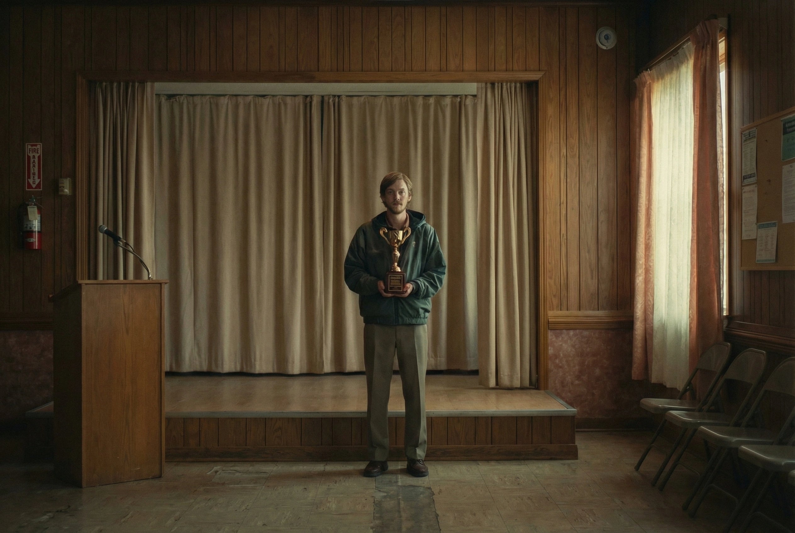 A young man stands alone in a wood-panelled community hall holding a small trophy. An empty podium stands to his left. Folding chairs line the wall to his right. The curtain behind him is closed. The room is empty. The trophy is his. The occasion is not entirely clear. The film  will explain everything.